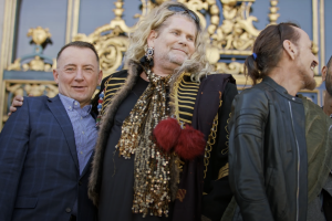 A person with long blonde hair and a gold-adorned outfit is embracing a person wearing a suit. They are standing in front of a building with gold detail on the front