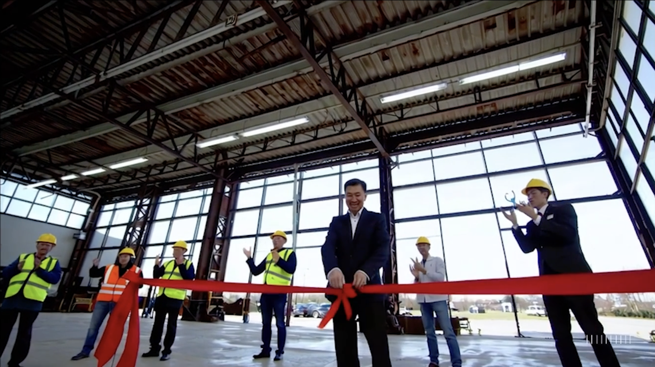 An AI-generated video of a factory floor with glass walls and a man in a black suit holding a floating red ribbon.