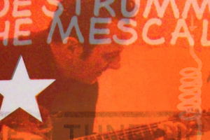A White man dressed in black leans over a guitar and strums. 