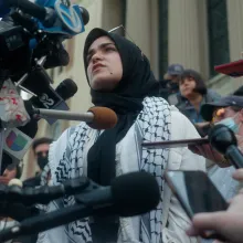 A young woman wearing a head scarf and a keffiyeh patterned scarf stands in the middle of a crowd of reporters holding microphones to her face.