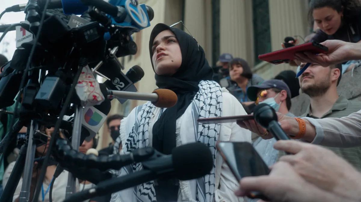 A young woman wearing a head scarf and a keffiyeh patterned scarf stands in the middle of a crowd of reporters holding microphones to her face.