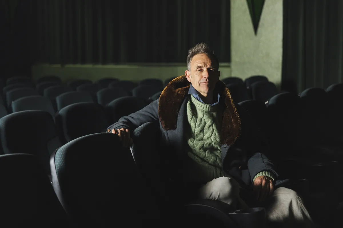 A white man sits in a darkened theater with a blue coat and green sweater.