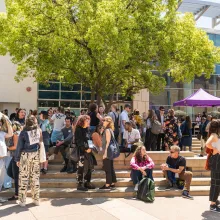 A crowd gathers outside a building. There is a tree with green leaves in the middle of the picture behind the crowd and in front of the building.