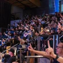 Audience members hold up their pointer finger, looking towards the stage offscreen.