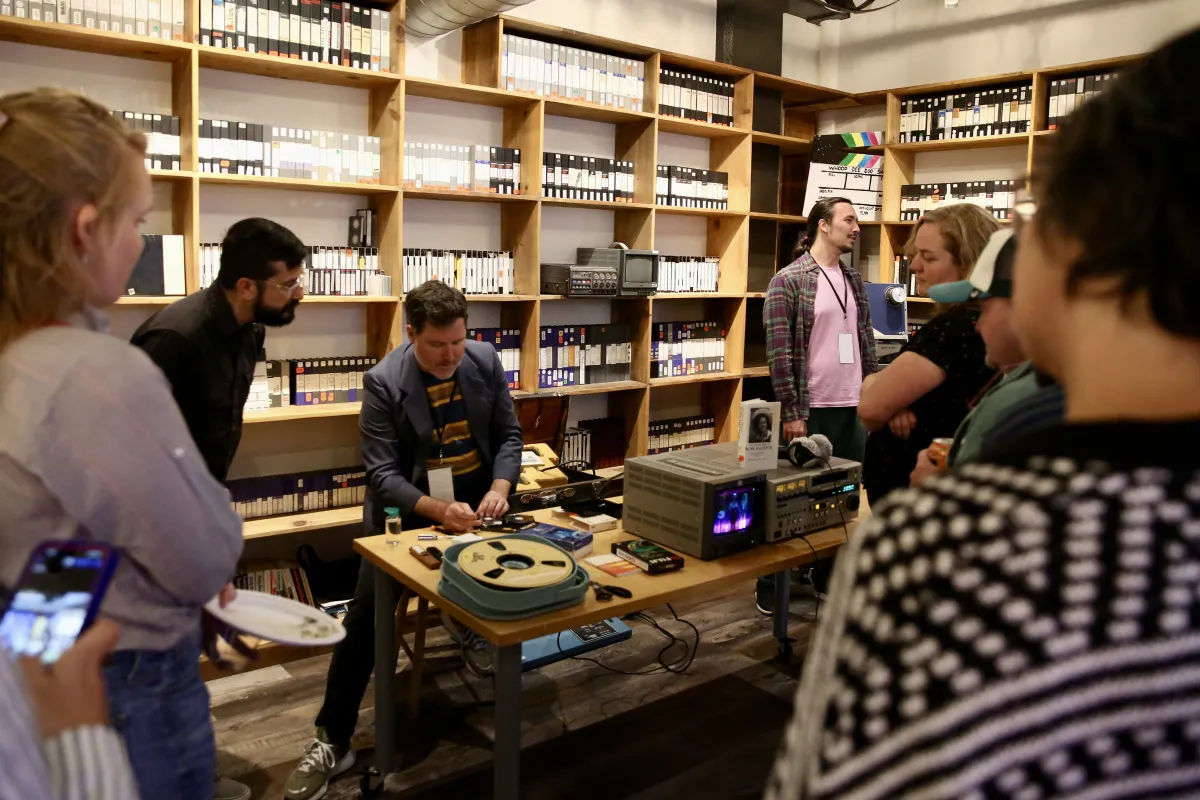 In a room with shelves lined with tapes, a group of people gather around a table with film materials.