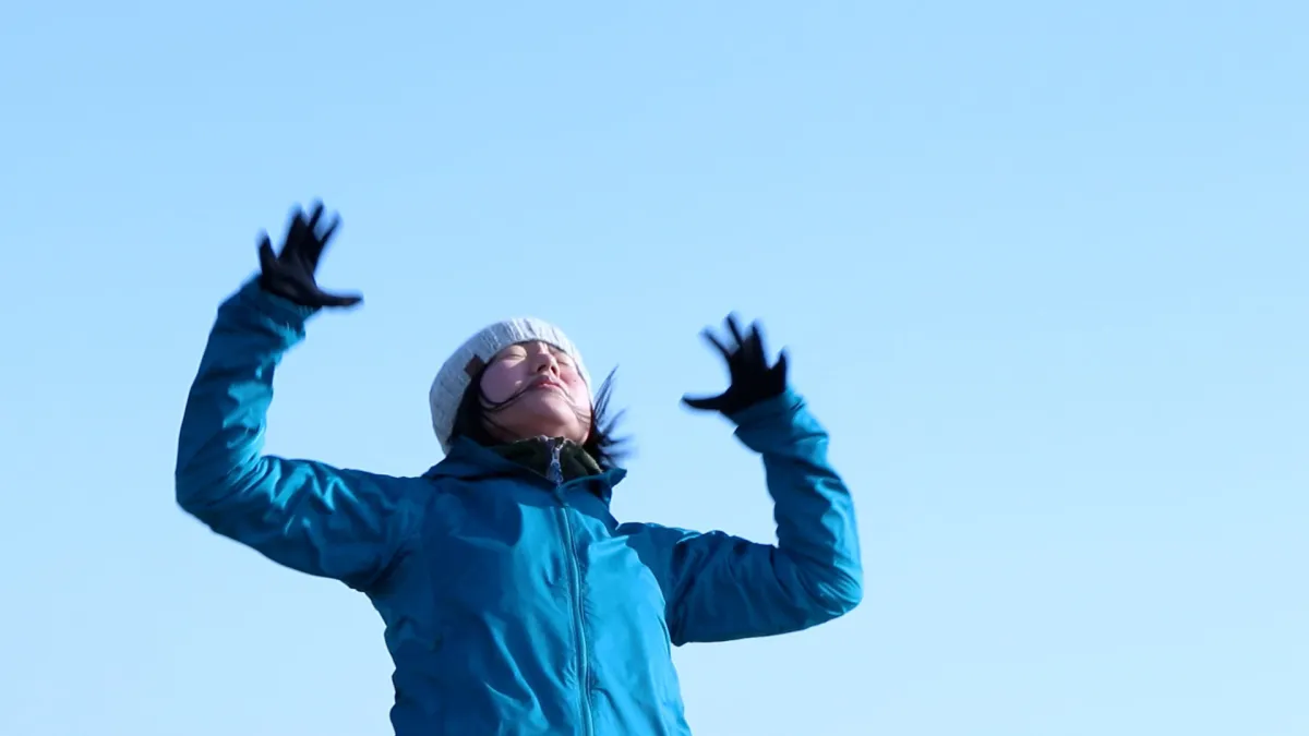 From below, a young Japanese woman wearing a beanie and a blue winter coat standards with arms outstretched.
