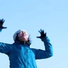 From below, a young Japanese woman wearing a beanie and a blue winter coat standards with arms outstretched.
