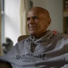 Harry Belafonte is a Black man, bald, smiling, wearing a grey hoodie and sitting on a chair, in front of a bookshelf. From Elvis Mitchell’s ‘Is That Black Enough For You?!?’ Courtesy of netflix