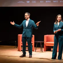 Two white people in navy blue suits stand on stage.