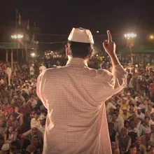 A man with brown skin in a white checkered shirt and white hat standing in front of a crowd. Still from 'An Insignificant Man' by Khushboo Ranka & Vinay Shukla. Courtesy of IFFLA. 