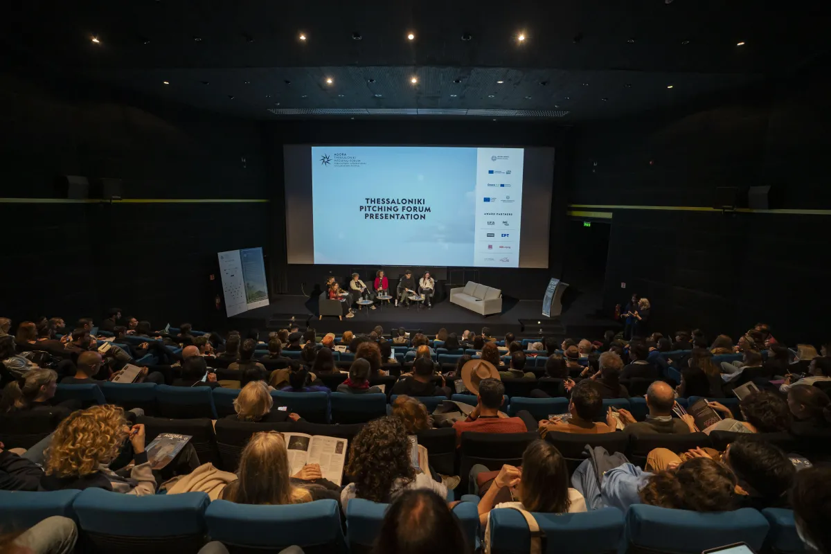 In a full auditorium, a group of panelists sit on coaches in front of a screen that reads: "Thessaloniki Pitching Forum Presentation".
