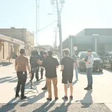 A group of white male filmmakers prep for a shot in a parking lot.