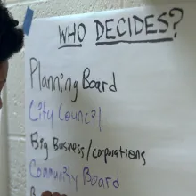 A black woman writes on a piece of large paper taped to a wall. On it is written: "Who Decides?"