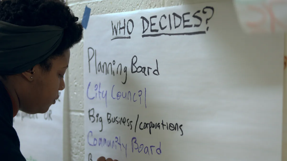 A black woman writes on a piece of large paper taped to a wall. On it is written: "Who Decides?"