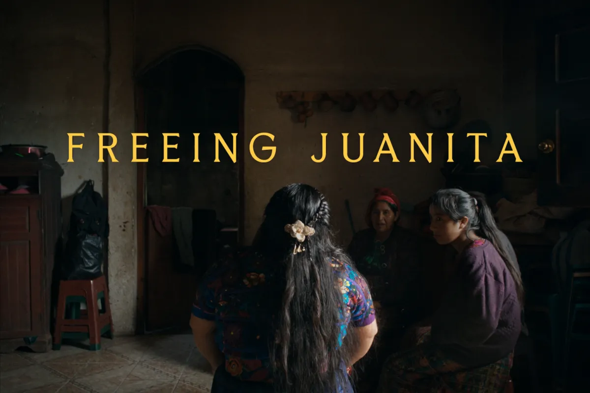 A woman from the back, sitting in front of other women, young and old, in a dark room. The woman in the center of the image has her hair braided any tied back with a white flower broche. She's wearing a colorful embroidered blue Maya huipil.