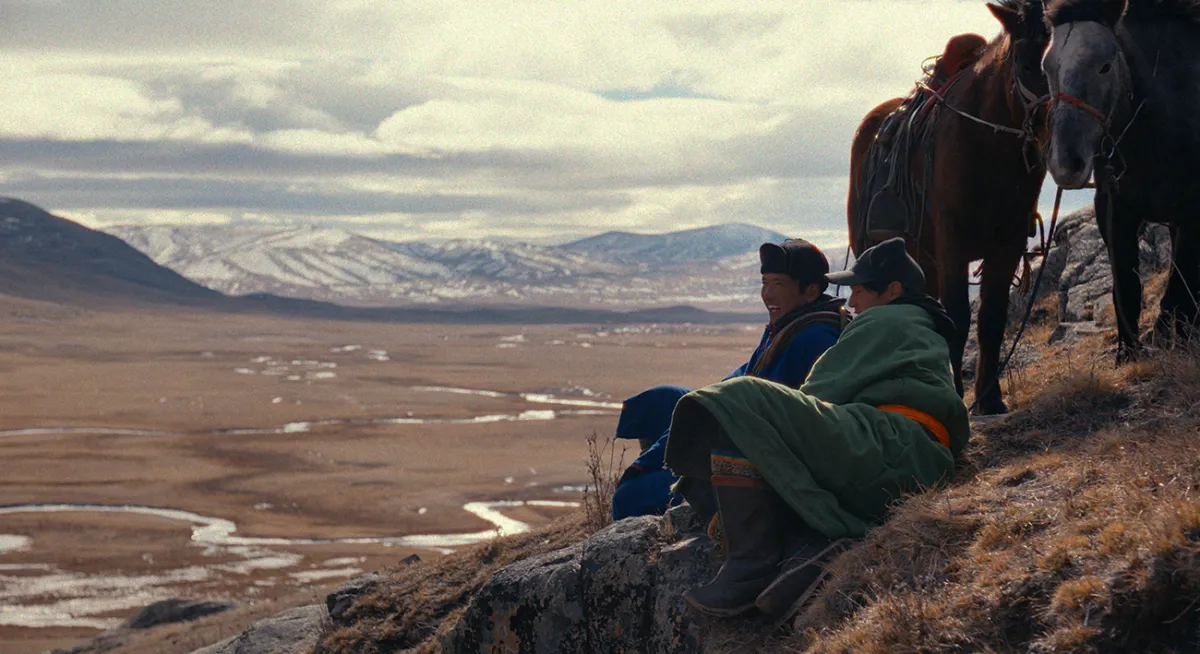 On a steep mountainside, two men sit on the ground next to their horses.