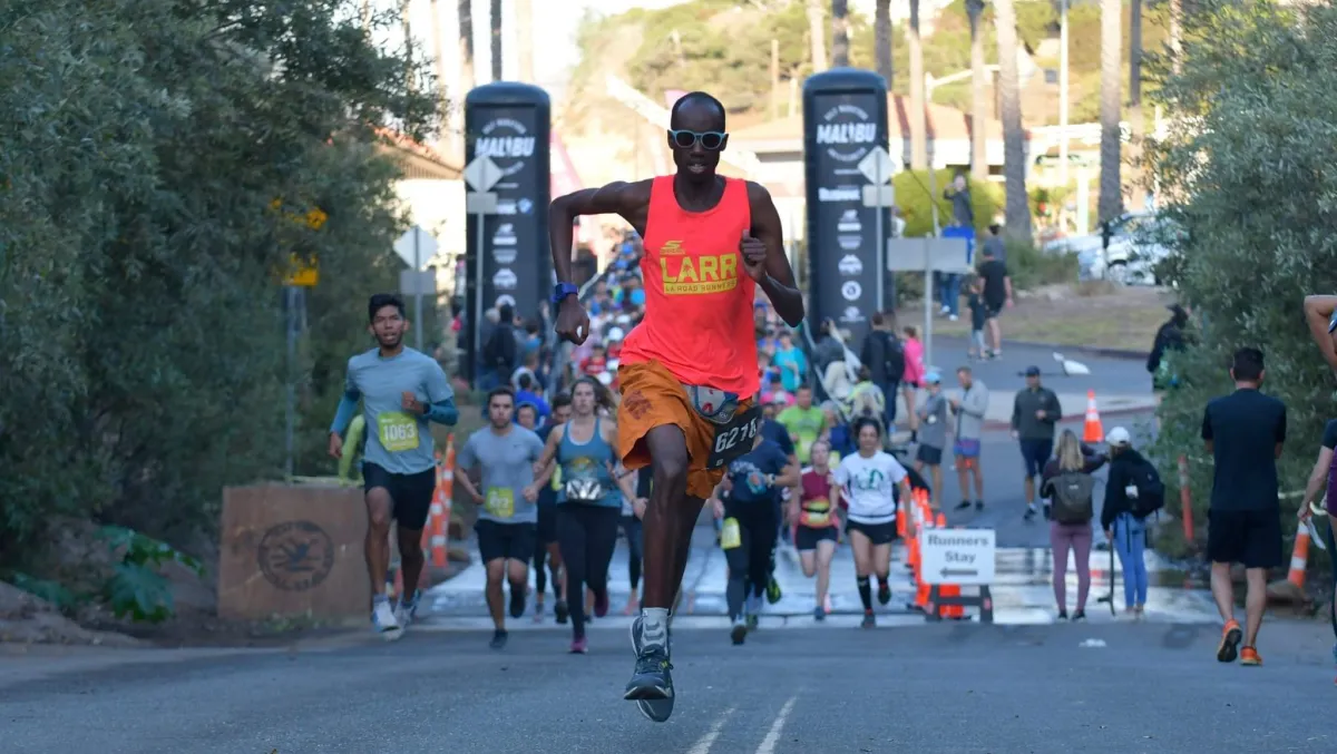 A man wearing sunglasses, shorts, and a neon colored top sprints ahead of a group of runners on a paved street