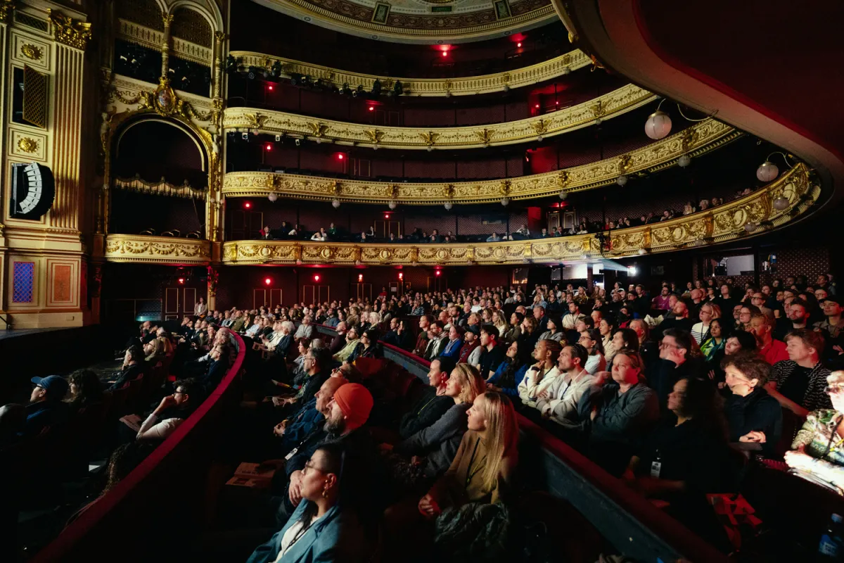 In a grand multi-story theater, a packed audience looks at an off-screen movie screen.