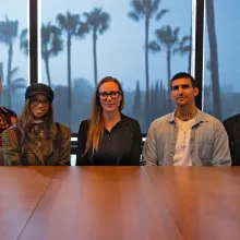 At a conference table, five people in laid back clothing smile at the camera. There are palm trees in the window behind them.