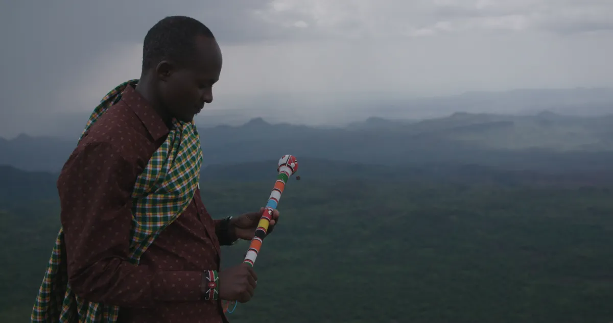 Black man holds a beaded staff and looks over a lush landscape
