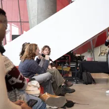In a bright room with concrete pillars and floors, a man in a sweater answers a question from the audience.