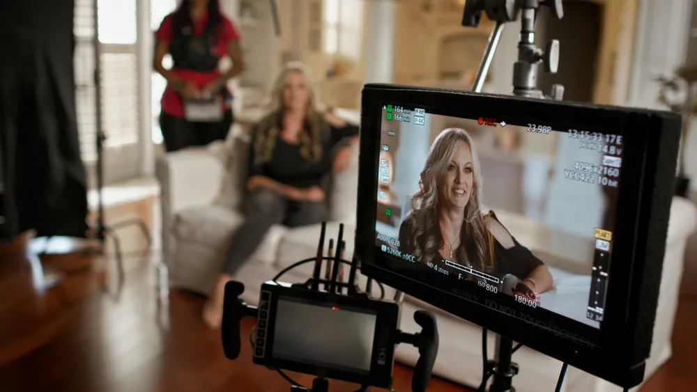 Stormy Daniels sitting in the background on a white armchair, with a monitor showing her close-up shot in the foreground.