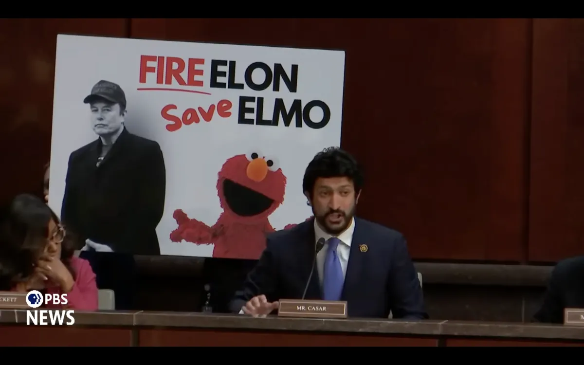 Pixelated screen shot of a U.S. congressman in front of a "Fire Elon Save Elmo" sign. The PBS News logo is in the bottom left corner.