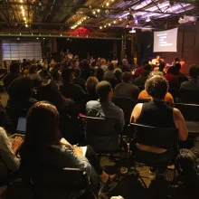 A crowded room of attendees sitting on fold-out chairs looks at a stage and presentation.