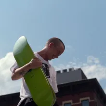 Leo Baker, a white person with buzz-cut hair, smiles toward the ground while holding a neon green skateboard deck. From Nicola Marsh and Giovanni Reda’s  ‘Stay on Board: The Leo Baker Story,. Photo courtesy of Netflix.