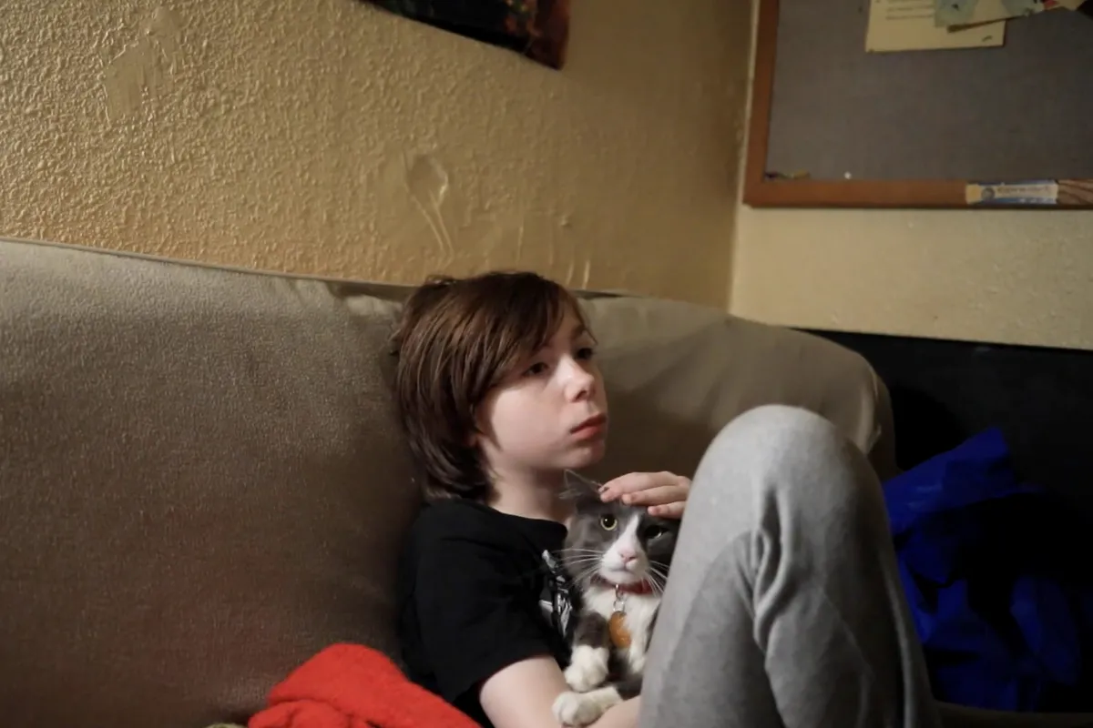 On a beige couch, a teenaged boy sits with his cat.